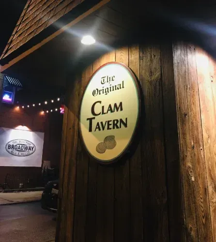 Oval wooden sign reading The Original Clam Tavern, lit at night on a wood-paneled wall