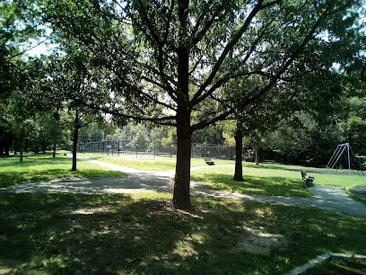 Shaded path and grass under large trees at Cobbs Creek Park, with benches, a fenced court and a swing set in the distance