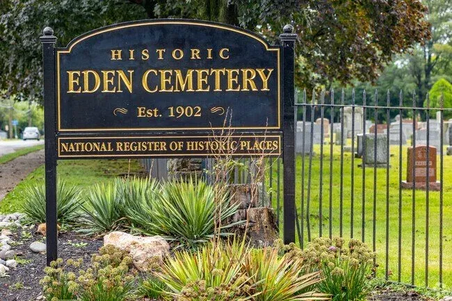 Black and gold Historic Eden Cemetery sign, established 1902, with National Register of Historic Places plaque beside an iron fence