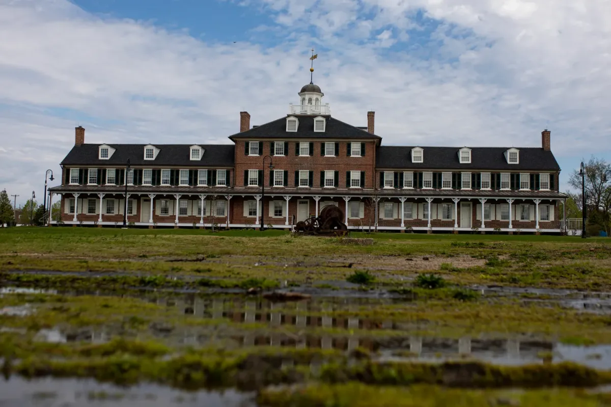 Red brick Lazaretto Quarantine Station building with a white cupola and long porch, seen across a grassy, puddled field